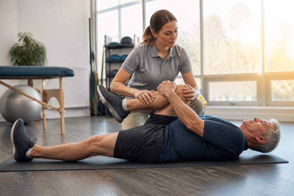 A physical therapist guiding a patient through a gentle lower back stretch on a mat in a bright clinic room, morning light through large windows, both wearing comfortable athletic clothing